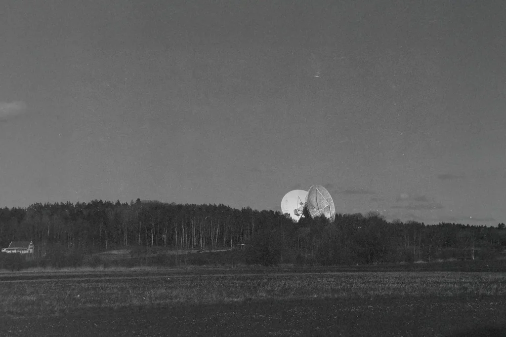 Large satellite dishes rising above a dense tree line in a wide, open field under a dark sky