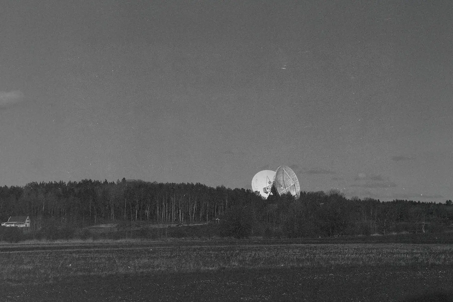 Large satellite dishes rising above a dense tree line in a wide, open field under a dark sky
