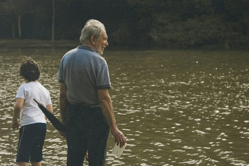 Older adult and child standing at the edge of a calm, reflective body of water, holding items in their hands while looking out across the surface