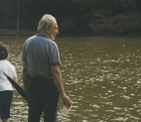 Older adult and child standing at the edge of a calm, reflective body of water, holding items in their hands while looking out across the surface