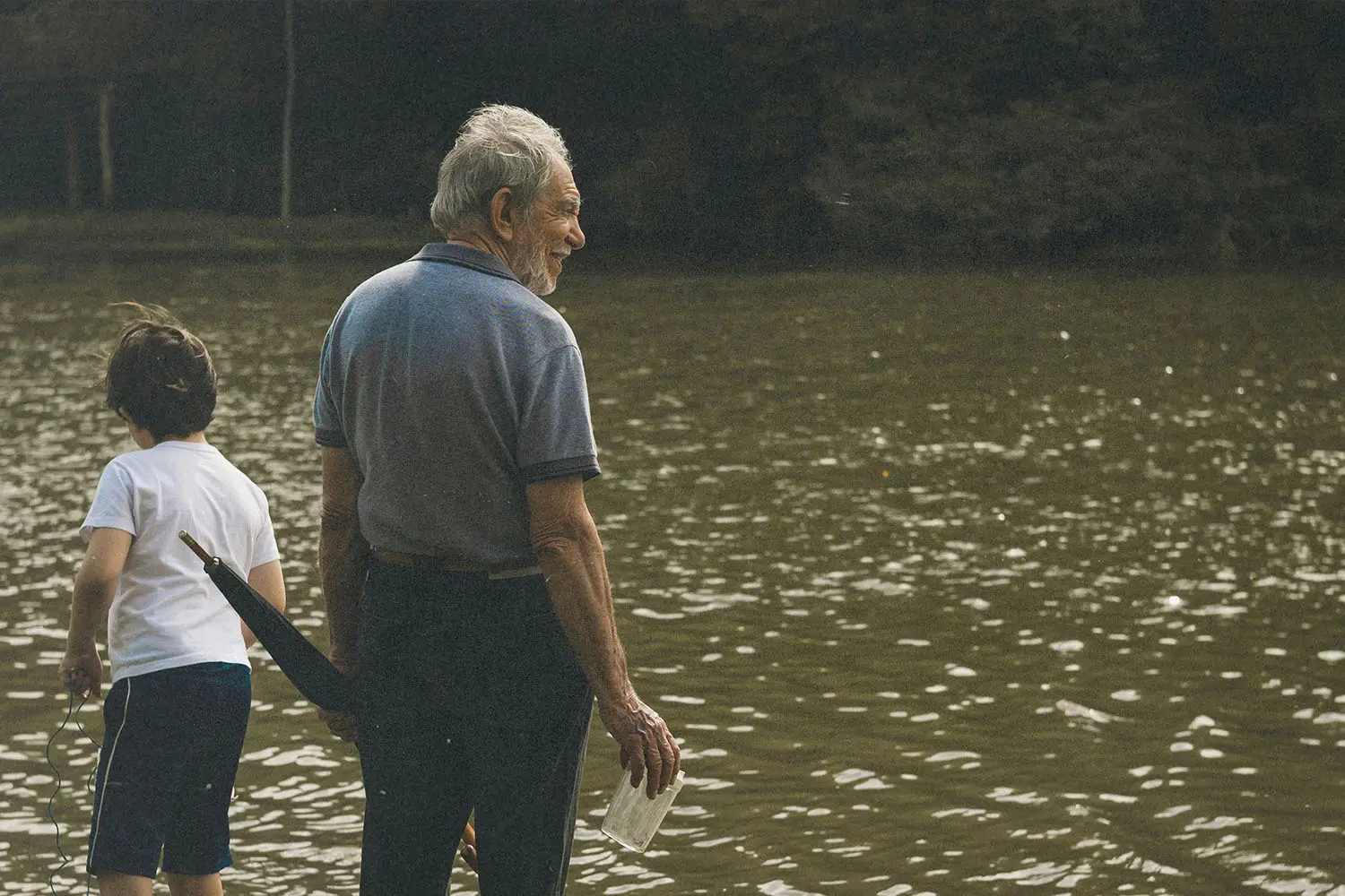 Older adult and child standing at the edge of a calm, reflective body of water, holding items in their hands while looking out across the surface