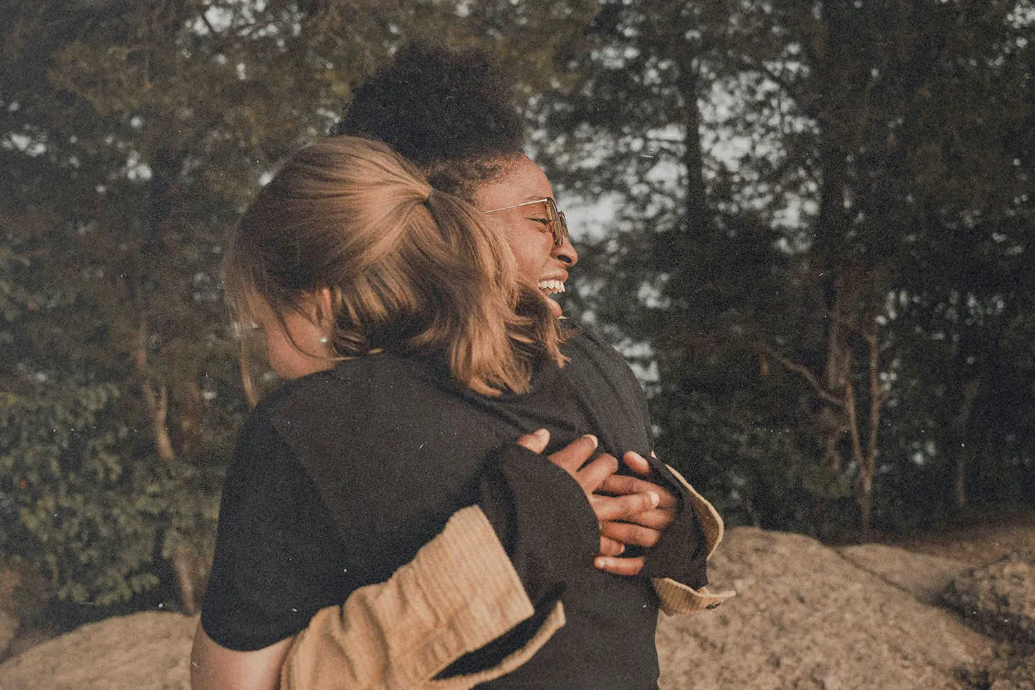 Two people embracing outdoors near large rocks, surrounded by trees and natural scenery