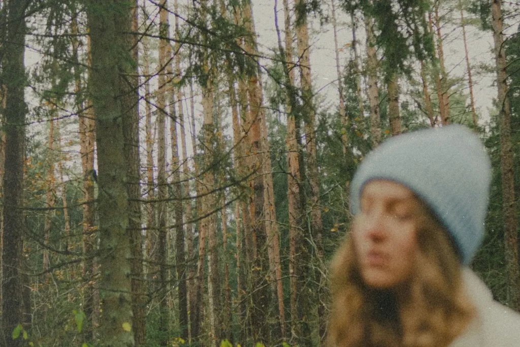Woman standing in a dense forest of tall pine trees wearing a knit winter hat