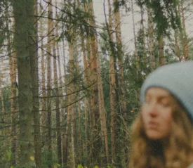 Woman standing in a dense forest of tall pine trees wearing a knit winter hat