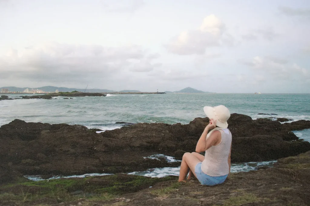 Woman wearing a wide-brimmed hat seated on seaside rocks facing the distant water
