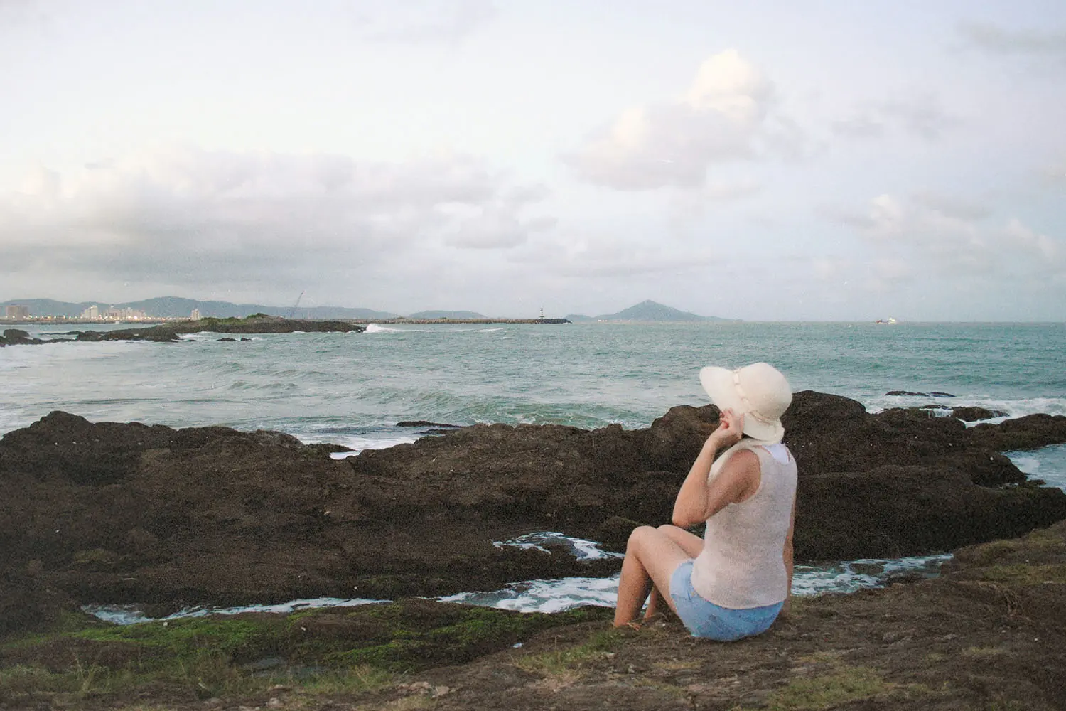 Woman wearing a wide-brimmed hat seated on seaside rocks facing the distant water