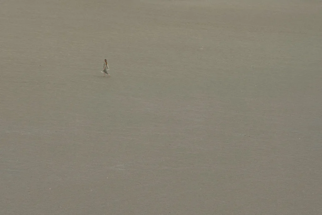 A single person walks across a vast, empty expanse of smooth sand, appearing small against the wide open landscape