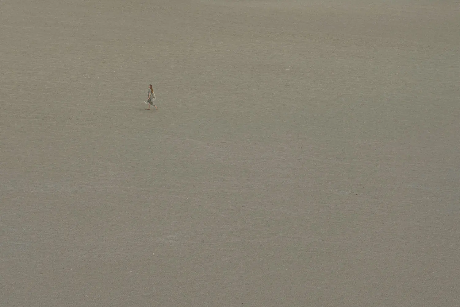 A single person walks across a vast, empty expanse of smooth sand, appearing small against the wide open landscape