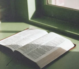 An open Bible rests on a windowsill, lit by soft natural light streaming through a nearby window