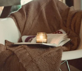 Cozy reading nook with a lit candle resting on an open book, draped with a chunky brown knit blanket over a white chair