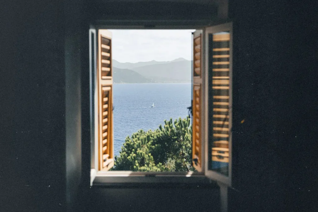 A window with wooden shutters opens to a scenic view of blue water, green treetops, and distant mountains