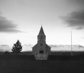 Small country church with a steeple standing before snow‑covered mountains at dusk in a black‑and‑white landscape