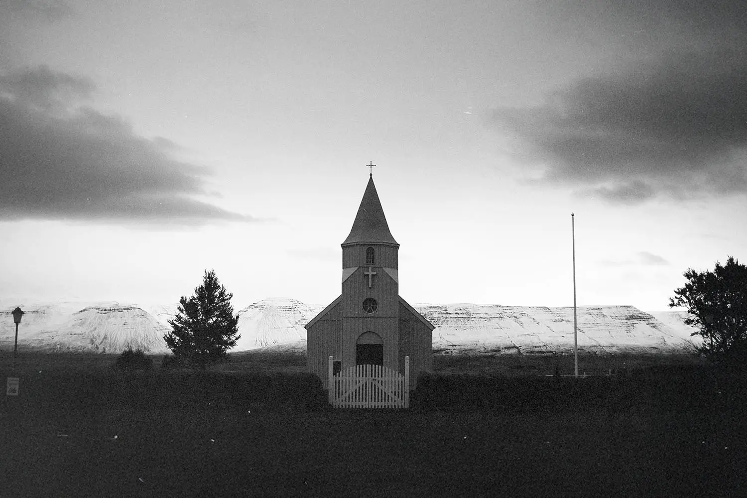 Small country church with a steeple standing before snow‑covered mountains at dusk in a black‑and‑white landscape