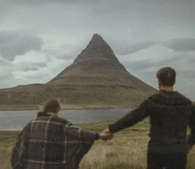Two people hold hands while facing a tall, cone‑shaped mountain beside a calm body of water