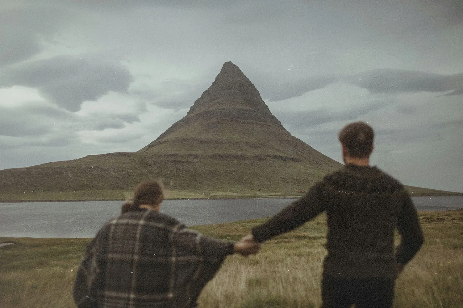 Two people hold hands while facing a tall, cone‑shaped mountain beside a calm body of water