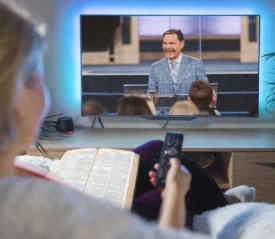 Woman sitting on the couch with a remote in her hand watching Kenneth Copeland on the TV