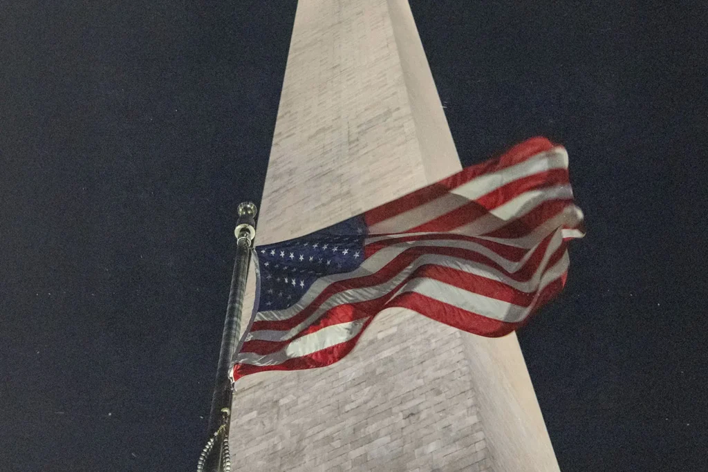An American flag waves at night in front of a tall stone monument, illuminated from below