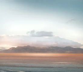 A distant mountain range sits beneath soft clouds as warm light spreads across a wide, flat desert foreground