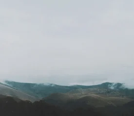 A series of low, rolling mountains are partially covered in mist beneath a gray, overcast sky