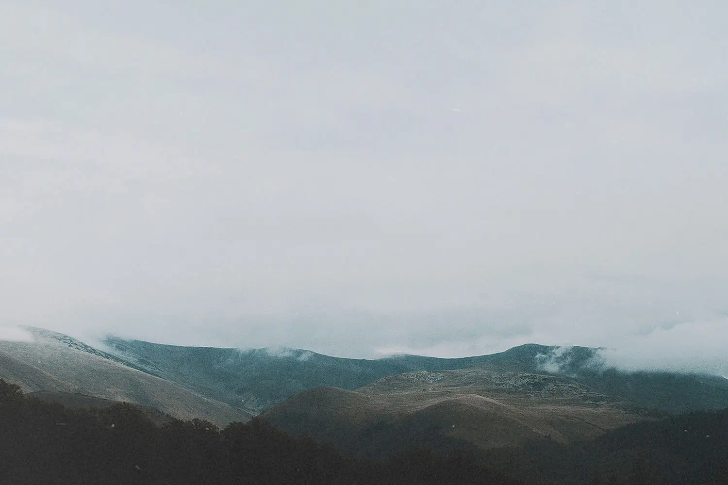 A series of low, rolling mountains are partially covered in mist beneath a gray, overcast sky