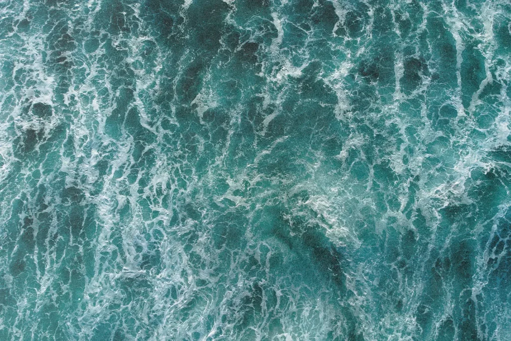 A top‑down view of churning ocean water shows swirling turquoise waves and white foam patterns