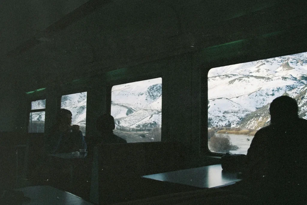 Passengers sit inside a dimly lit train car, looking out wide windows at snow‑covered mountains and a winding river