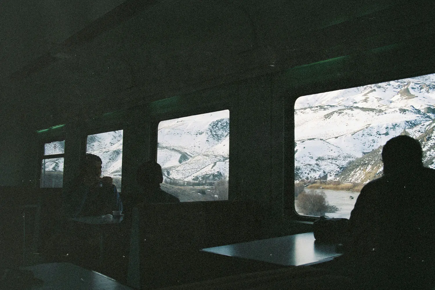 Passengers sit inside a dimly lit train car, looking out wide windows at snow‑covered mountains and a winding river