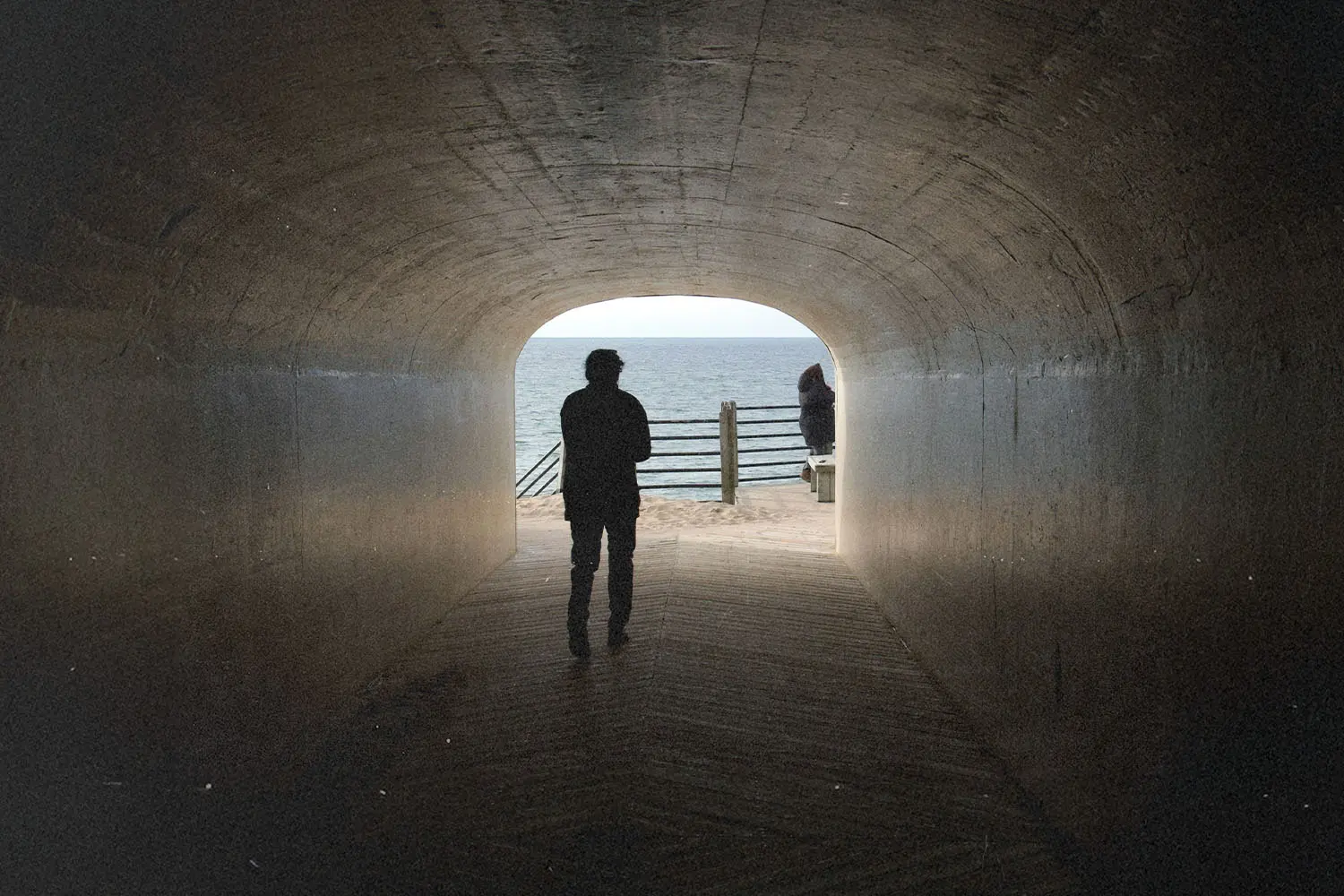 Silhouetted person walking through a dark tunnel toward a bright ocean view, with another figure standing near the railing outside