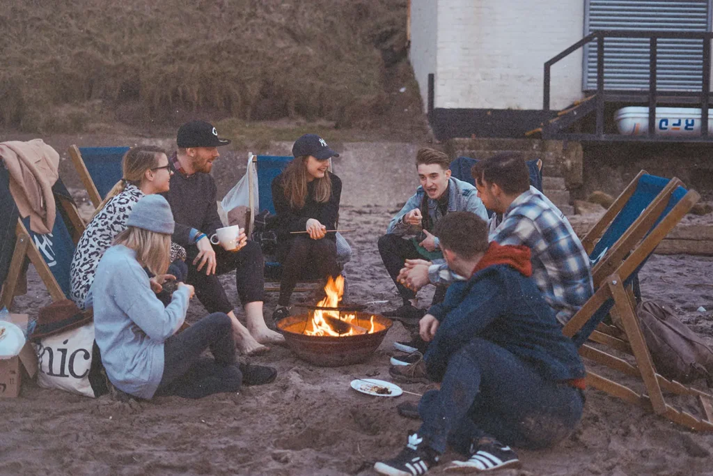 Group of friends sitting around a small beach bonfire, chatting and relaxing in deck chairs