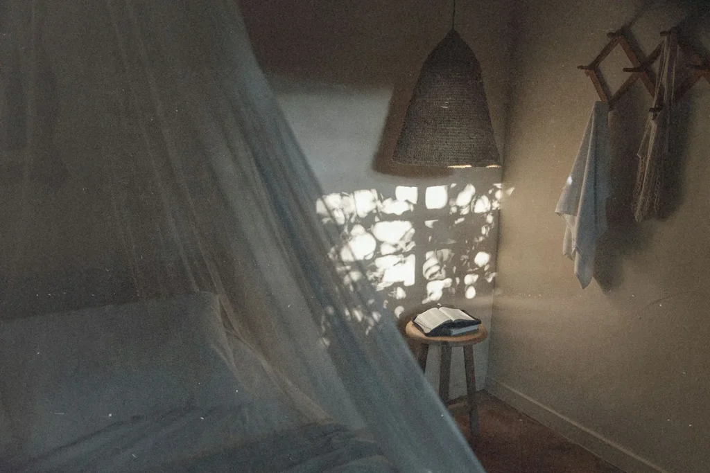 Soft morning light filters through a window, casting patterned shadows onto a small table holding an open book in a calm bedroom