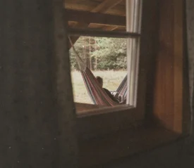 Person relaxing in a striped hammock under a wooden porch, viewed through a window looking out toward a grassy area and trees