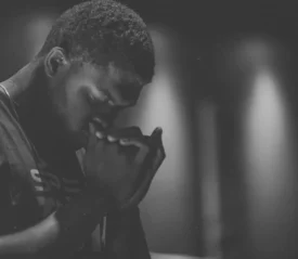 Person standing in a dimly lit room with hands raised near their face in a posture of reflection or prayer