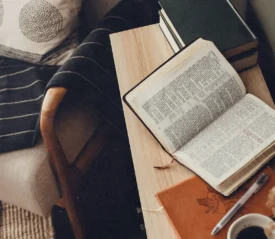 Open Bible rests on a wooden side table beside a journal, pen, stacked books, and a cup of coffee near a cozy chair