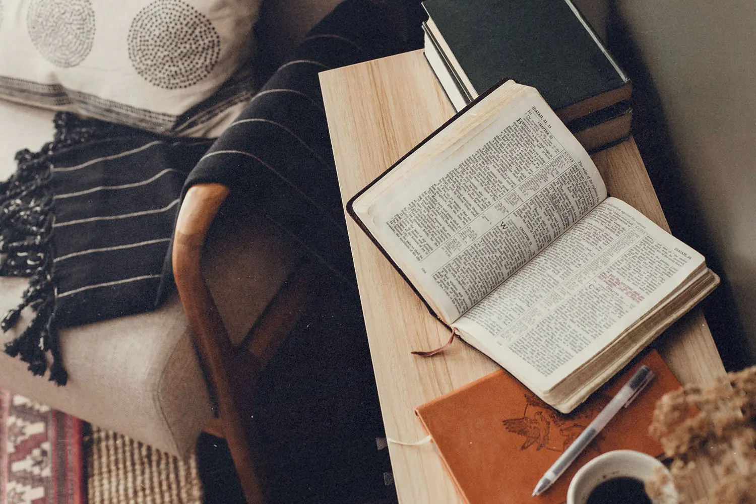 Open Bible rests on a wooden side table beside a journal, pen, stacked books, and a cup of coffee near a cozy chair