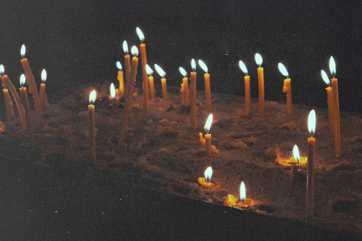 Dozens of thin, glowing candles stand upright in sand, their flames flickering softly in the dark