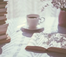 Open book lying on a sunlit table beside a stack of books, a cup of coffee, and a vase of dried flowers with soft shadows cast across the surface