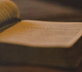 Close-up of an open Bible lying on a table, softly lit with warm, golden light highlighting the printed text