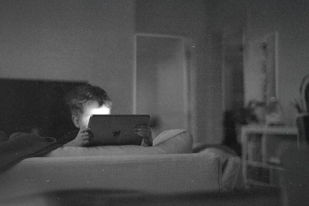 A child sits on a couch in a dim room, holding a tablet with soft light reflecting from the screen