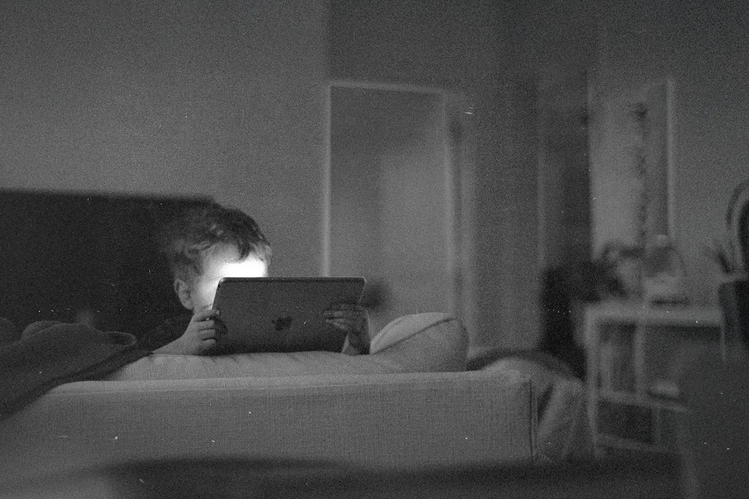 A child sits on a couch in a dim room, holding a tablet with soft light reflecting from the screen