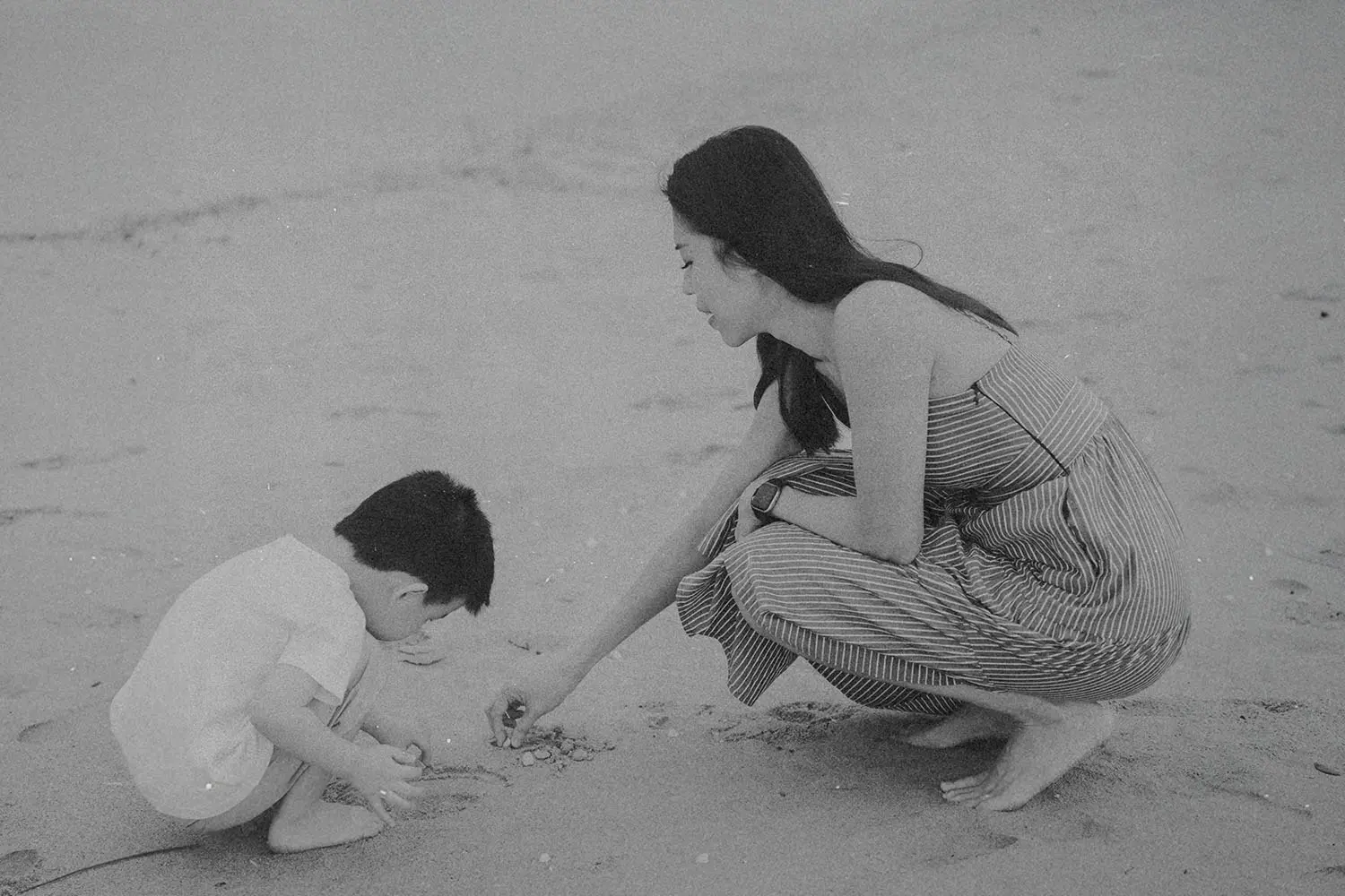 A woman kneels on the beach beside a child as they look closely at small objects in the sand