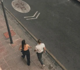 Two people walk side by side on a city sidewalk, viewed from above as they move along the street