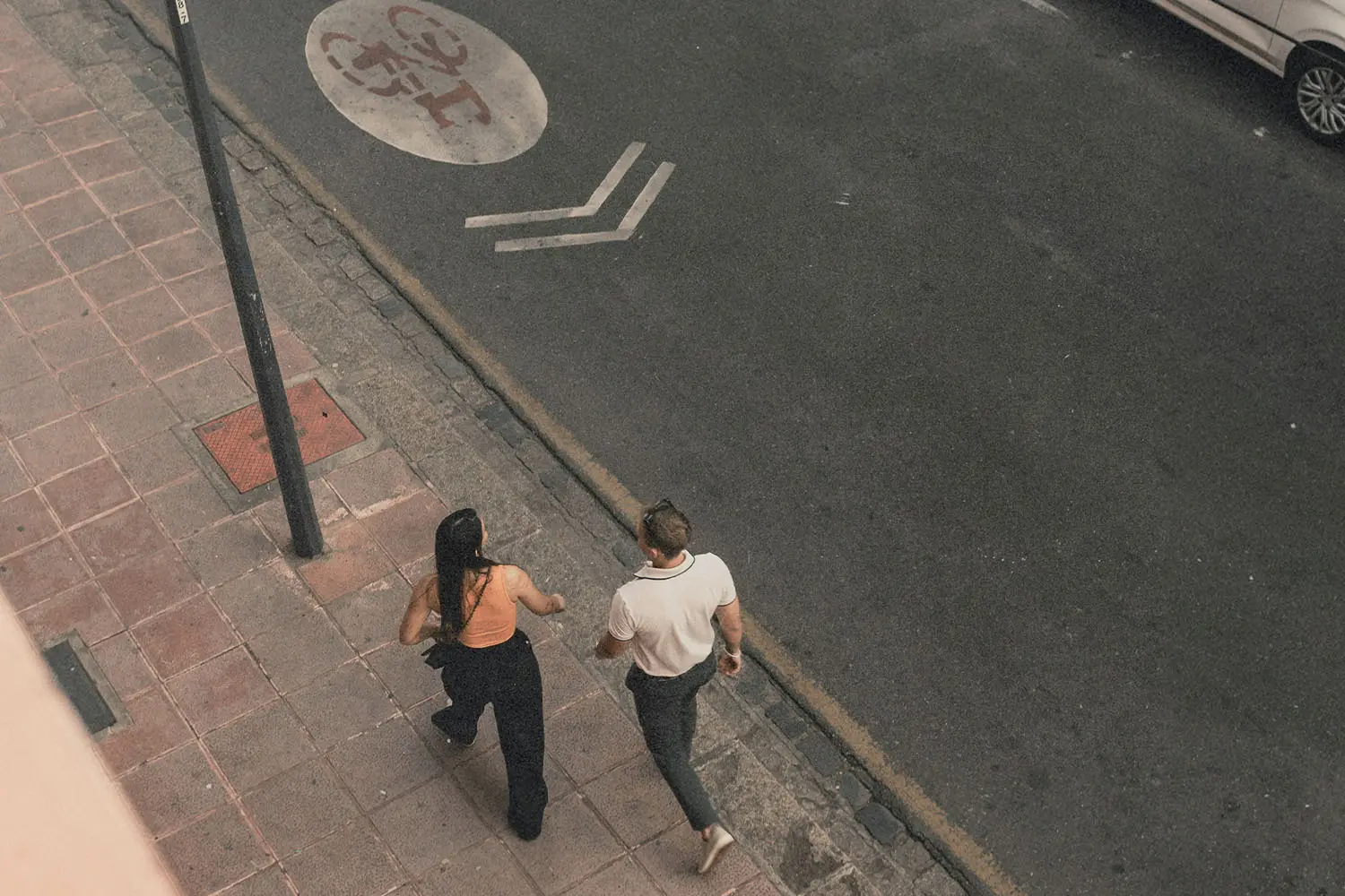Two people walk side by side on a city sidewalk, viewed from above as they move along the street