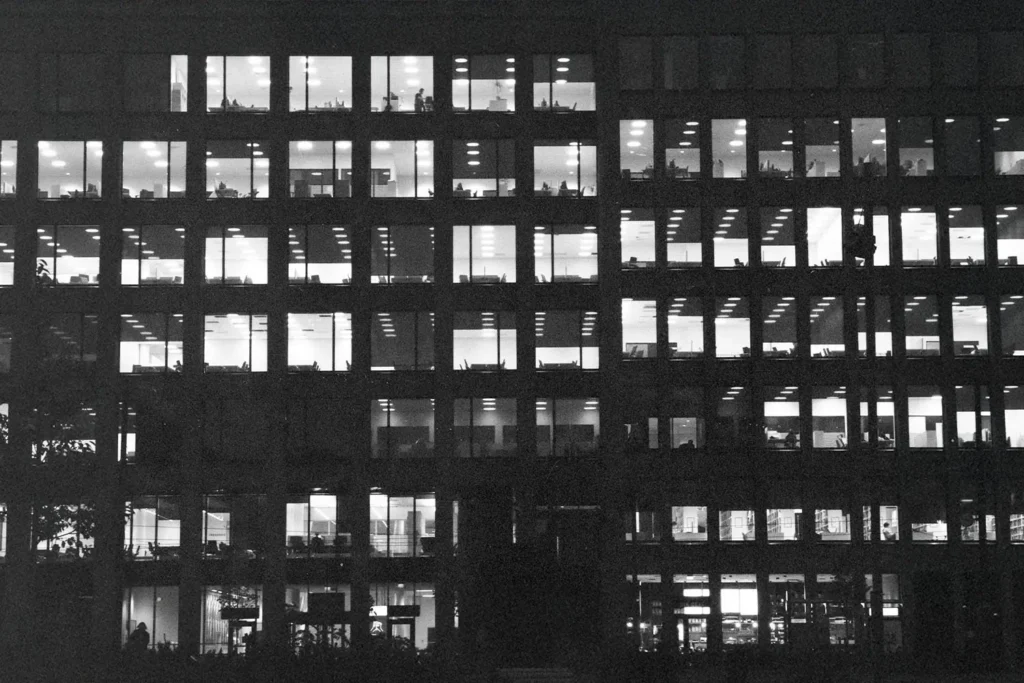 A nighttime view of a large office building, its grid of windows illuminated to reveal people working inside