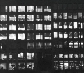 A nighttime view of a large office building, its grid of windows illuminated to reveal people working inside