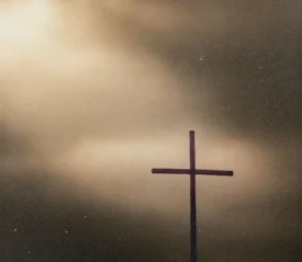 A simple wooden cross stands silhouetted against a dramatic sky with a beam of light breaking through the clouds