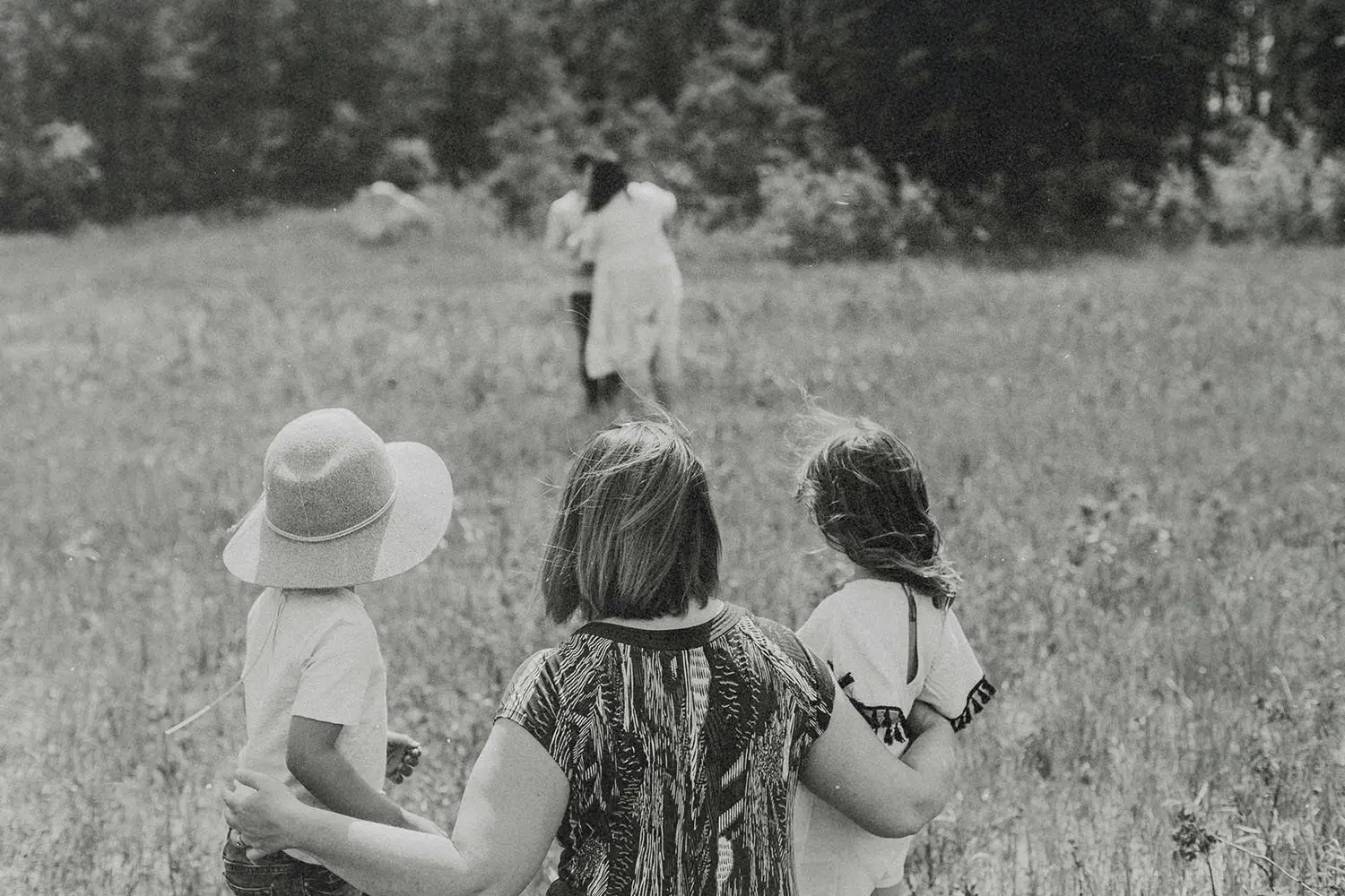 A woman stands in a grassy field with two young children at her sides, all facing toward another pair of people walking ahead across the open meadow, with trees lining the distance