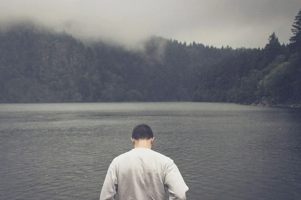 A person stands at the edge of a calm lake surrounded by misty, forested mountains