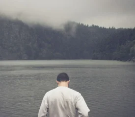A person stands at the edge of a calm lake surrounded by misty, forested mountains