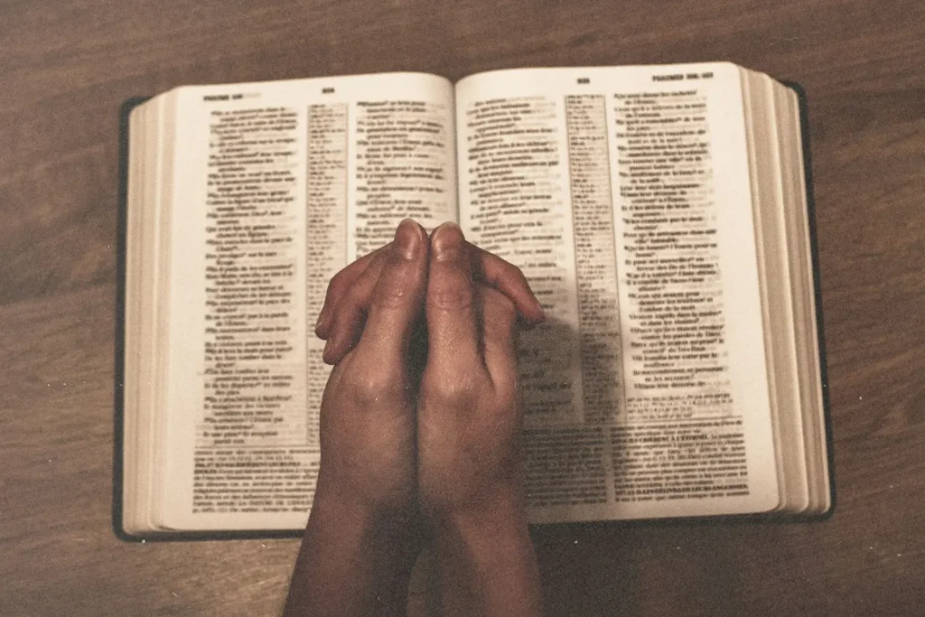 Hands folded in prayer over an open Bible on a wooden surface