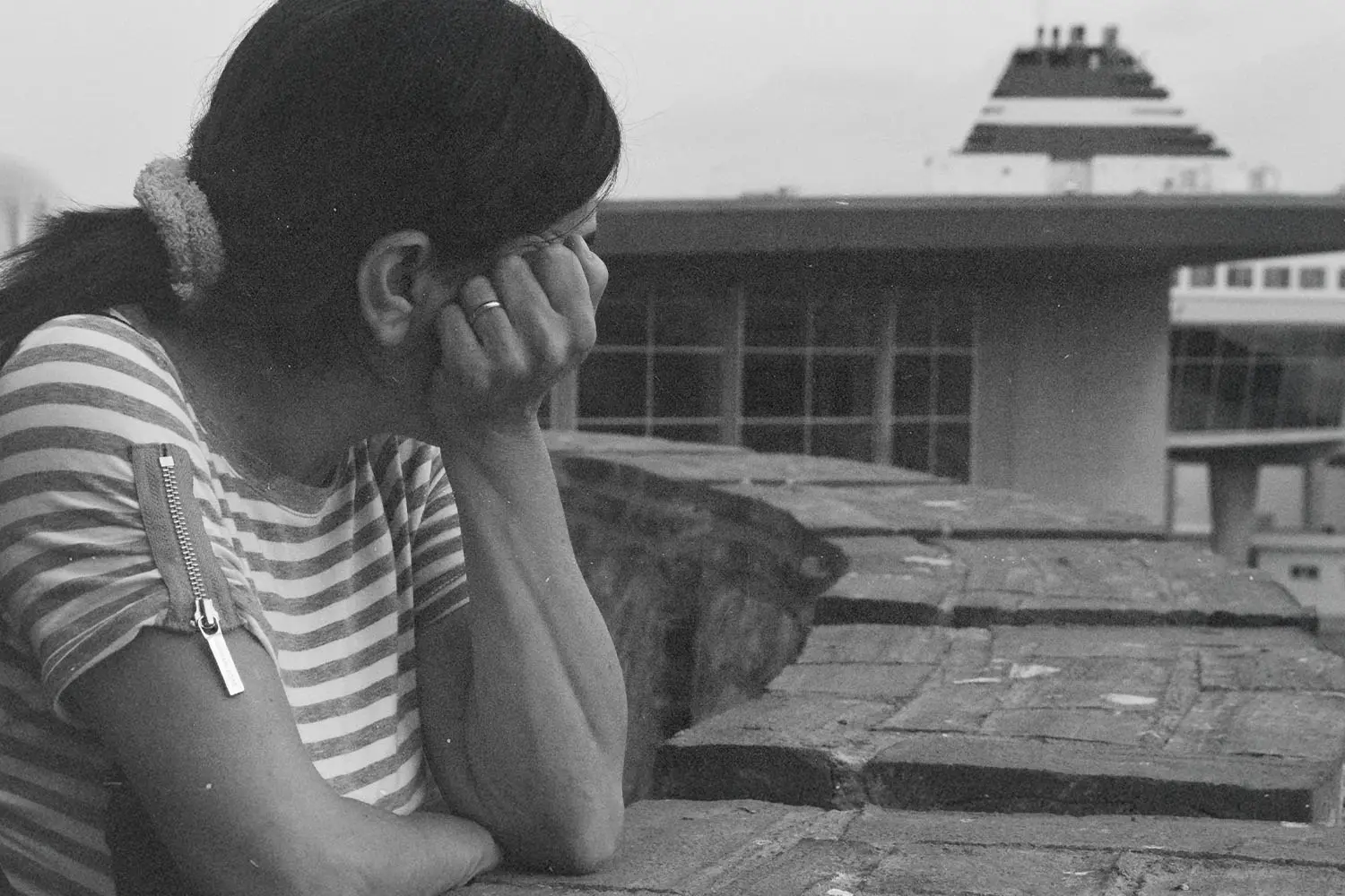 A person wearing a striped shirt leans on a brick wall, looking out toward a large docked ship in the distance